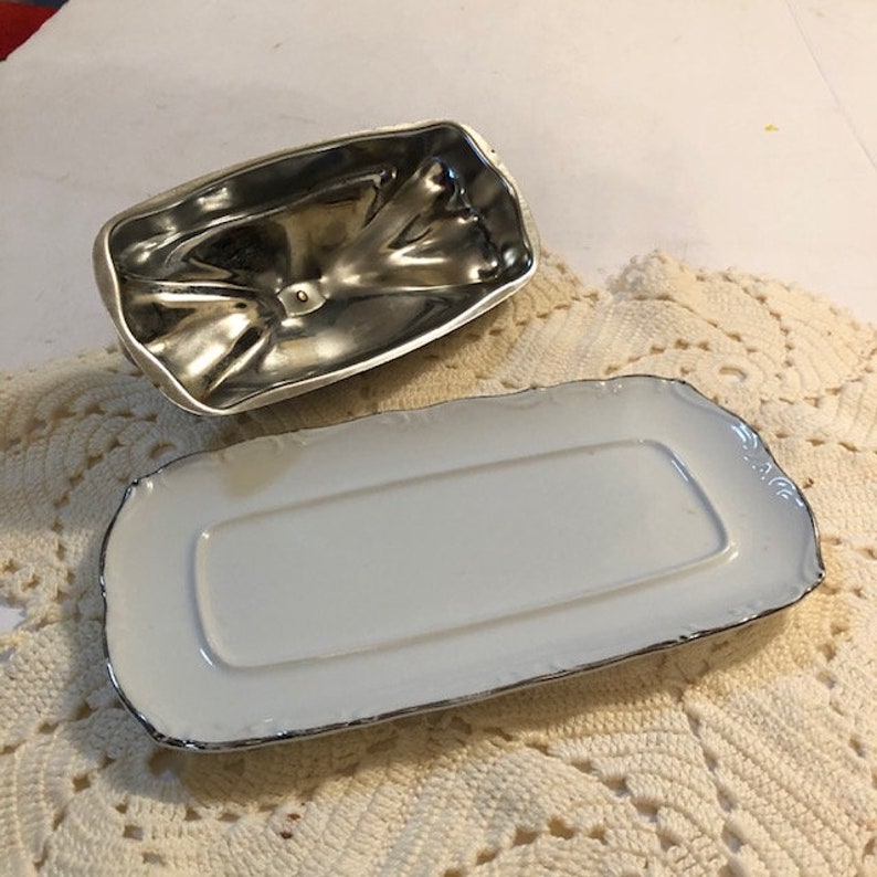 May include: A white porcelain butter dish with a silver-plated lid. The dish has a scalloped edge and a silver trim. The lid has a decorative design.