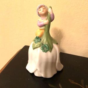 May include: A porcelain figurine of a child holding a green stem, atop a white flower-shaped base. The child wears a lavender hat, pink sleeves, and yellow clothing. The figurine is a decorative item.