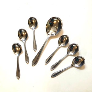 May include: Set of seven silver spoons, including a larger serving spoon. The spoons have a simple design with a rounded bowl and a slightly curved handle.