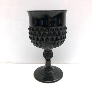 May include: A black glass goblet featuring a textured diamond point pattern on the bowl and a matching geometric design on the stem. The piece has a smooth, flared rim, a slender stem, and a wide, circular base, all crafted from solid black glass.