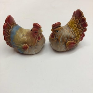 May include: A pair of ceramic chicken-shaped salt and pepper shakers. The left chicken features a tan body with blue and red wing accents. The right chicken has a tan body with yellow and red feather details. Both have red combs and wattles.