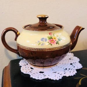 May include: A vintage teapot with a brown base and lid, and a cream-colored center adorned with a floral design. The teapot has a gold trim and handle, and sits on a white lace doily.