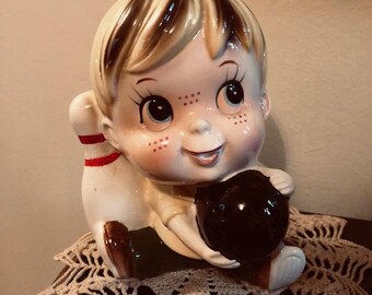 May include: A ceramic figurine of a boy holding a bowling ball and a bowling pin. The boy has blonde hair, brown eyes, and freckles. He is wearing a green shirt and brown pants.