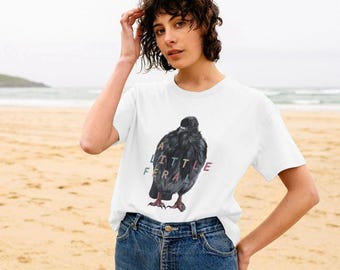 Puede incluir: Una camiseta blanca de manga corta con un gráfico de una paloma gris oscuro en el pecho. El texto "A LITTLE FERAL" está impreso en letras multicolores sobre la ilustración del ave. La camiseta se combina con unos vaqueros de mezclilla azul.