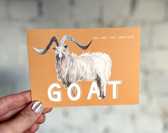 Puede incluir: Tarjeta de felicitación con una ilustración de una cabra con cuernos grandes y pelaje blanco. La palabra "GOAT" está impresa en letras blancas grandes debajo de la cabra. El texto "YOU ARE THE ABSOLUTE" está impreso encima de la cabra.