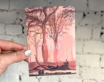 Puede incluir: Una tarjeta rectangular con una pintura de acuarela de una escena de bosque en tonos marrones y rosas. Las palabras "LOVE YOU" están impresas en blanco. La tarjeta se sostiene contra una pared de ladrillos blancos.
