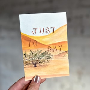 May include: A greeting card with a watercolor desert scene. The card features the words "JUST TO SAY" in brown lettering. The artwork depicts a desert landscape with sand dunes, a small tree, and a light blue sky.