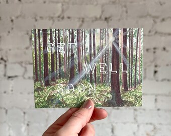 Puede incluir: Una tarjeta rectangular de "Get Well Soon" con una escena de bosque con árboles altos y luz solar. La tarjeta tiene un fondo blanco con el texto en una fuente blanca y elegante. La imagen es sostenida por una persona.