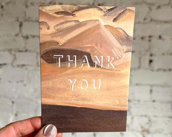 Puede incluir: Una tarjeta de felicitación rectangular con un diseño de paisaje en acuarela en tonos marrones y beige. Las palabras "THANK YOU" están impresas en blanco, con una fuente sans-serif en el centro de la tarjeta. La parte inferior de la tarjeta es marrón oscuro.