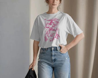 Puede incluir: Camiseta blanca con un diseño de flamenco rosa. El diseño presenta varios flamencos, plantas y elementos tropicales. La camiseta se combina con vaqueros azul claro.