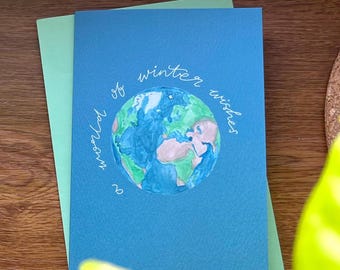 Puede incluir: Tarjeta de felicitación azul con una ilustración en acuarela de la Tierra, rodeada por las palabras "world of winter wishes" en escritura blanca. La tarjeta está sobre una tarjeta verde claro, con un fondo de madera.