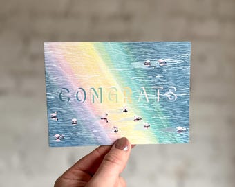 Puede incluir: Una tarjeta de felicitación con la palabra "CONGRATS" en blanco, sobre un fondo de acuarela. El fondo presenta un degradado de arcoíris y una escena de agua azul con pequeños pájaros blancos. La tarjeta es sostenida por una mano.