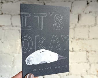 Puede incluir: Una tarjeta de felicitación gris oscuro con las palabras "IT'S OKAY" en letras blancas delineadas. Una ilustración blanca de una foca está debajo del texto. La frase "WE ALL HAVE THOSE DAYS" está impresa en la parte inferior.