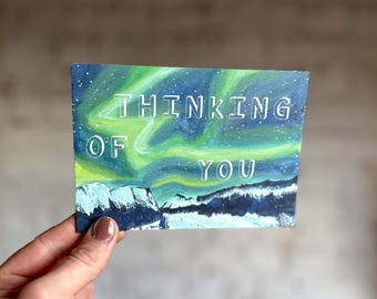 Puede incluir: Una tarjeta rectangular con una escena pintada de la Aurora Boreal. La obra de arte presenta un cielo verde y azul vibrante con las palabras "THINKING OF YOU" en letras blancas. Debajo, una cordillera.