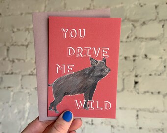 Puede incluir: Una tarjeta de felicitación con fondo rojo y el texto "YOU DRIVE ME WILD" en blanco. Una ilustración en negro y marrón de un jabalí está debajo del texto. La tarjeta está dentro de un sobre rosa.
