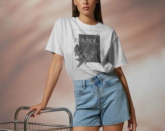 Puede incluir: Una camiseta blanca de manga corta con un gráfico rectangular que incluye el texto PISCES y una ilustración de dos peces. La prenda se combina con unos pantalones cortos de mezclilla azul claro de talle alto y diseño de cinco bolsillos.