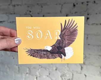 Puede incluir: Tarjeta de felicitación amarilla mostaza con la frase "YOU WILL SOAR" y una ilustración de un águila calva en vuelo. El águila tiene plumas marrones y blancas.