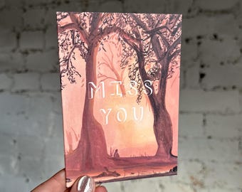 Puede incluir: Tarjeta rectangular con una pintura de acuarela de una escena de puesta de sol. Dos árboles enmarcan la puesta de sol, con las palabras "MISS YOU" en blanco. La tarjeta está sostenida por una mano con esmalte de uñas plateado.