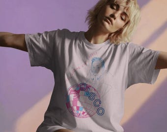 Puede incluir: Camiseta blanca con un diseño gráfico que presenta una bola de discoteca rosa y azul, la palabra "DISCO" y un dibujo lineal de una persona. También está presente el texto "I've been missing a little...". La camiseta la lleva una persona.