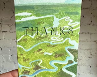 Puede incluir: Una tarjeta de felicitación de acuarela con un diseño de paisaje. La tarjeta presenta un paisaje verde y azul con la palabra "THANKS" en letras negras. La tarjeta se sostiene contra una pared de ladrillos blancos.