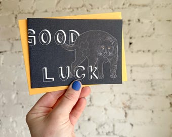 Puede incluir: Tarjeta de felicitación negra con las palabras blancas "GOOD LUCK" y una ilustración de pantera. La tarjeta está sobre un fondo amarillo.