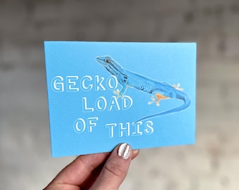 Puede incluir: Una tarjeta azul claro con el texto "GECKO LOAD OF THIS" y una ilustración de un gecko azul. El gecko tiene patas naranjas y está enroscado. La tarjeta se sostiene en una mano con esmalte de uñas plateado.