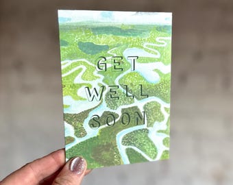 Puede incluir: Tarjeta de felicitación rectangular con las palabras "GET WELL SOON" en letras negras. La tarjeta presenta una pintura a la acuarela de un paisaje verde y blanco. La tarjeta está sujeta por una mano con esmalte de uñas brillante.