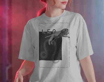 Puede incluir: Una camiseta gris claro de manga corta con un gráfico en blanco y negro de un león saltando sobre la palabra LEO. El diseño incluye un mapa de constelaciones, el símbolo del zodiaco Leo y varias palabras descriptivas en un marco.