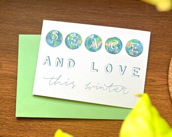 Puede incluir: Una tarjeta de felicitación con las palabras "PEACE AND LOVE" y "this winter" en una fuente manuscrita. La palabra "PEACE" está escrita con cada letra dentro de un globo terráqueo. La tarjeta está sobre un sobre verde.