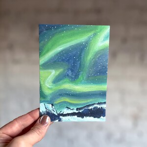 May include: A small rectangular painting depicts the aurora borealis. The artwork features a vibrant display of green and blue hues, with a starry night sky above snow-covered mountains. The painting is held by a hand.