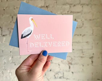 Puede incluir: Una tarjeta de felicitación rosa con una ilustración de cigüeña y la frase blanca "WELL DELIVERED". Se acompaña de un sobre azul. La cigüeña es blanca con detalles negros en las alas y un pico amarillo. La tarjeta se sostiene en una mano.