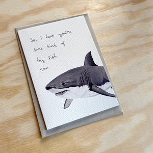 May include: A greeting card featuring a watercolor illustration of a shark. The shark is depicted in shades of gray and white, with visible teeth. The card includes handwritten text that reads, "So, I hear you're some kind of big fish now."