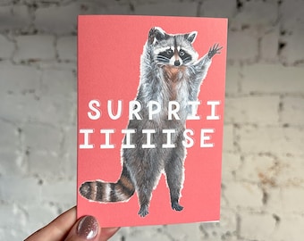 Puede incluir: Una tarjeta de felicitación con un fondo rojo que presenta una ilustración de un mapache. El mapache está de pie con un brazo levantado. La palabra "SURPRISE" está impresa en letras blancas en la parte superior de la tarjeta.