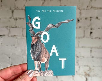 Puede incluir: Tarjeta de felicitación turquesa con una ilustración de una cabra Markhor y el texto "YOU ARE THE ABSOLUTE GOAT". La cabra tiene cuernos largos y en espiral y un pelaje marrón y blanco. La tarjeta está sujeta por una mano.