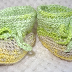 May include: A pair of crocheted baby booties in green and yellow. The booties have a lace-up design and are made with a soft yarn.