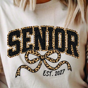 May include: A beige t-shirt featuring the word SENIOR in black glittery letters with a leopard print outline. Below the text is a large leopard print bow graphic and the black text EST. 2027. The shirt has short sleeves and a crew neckline.
