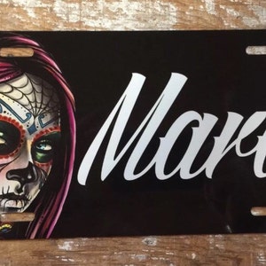 May include: A black and white personalized license plate with a colorful sugar skull design and the name "Mara" in white cursive lettering.