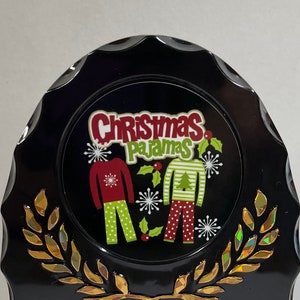 May include: Black and gold Christmas ornament with a white circle featuring the text "Christmas Pajamas" and a graphic of two people wearing red and green pajamas with snowflakes and holly.