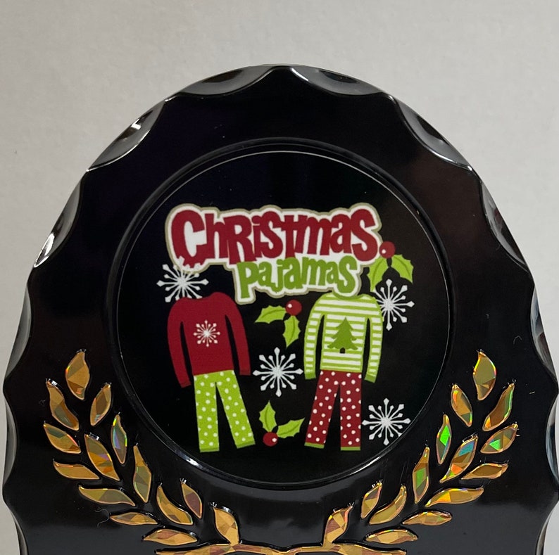 May include: Black and gold Christmas ornament with a white circle featuring the text "Christmas Pajamas" and a graphic of two people wearing red and green pajamas with snowflakes and holly.