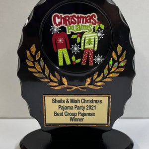May include: Black and gold award plaque with a Christmas pajama party theme. The plaque features a circular image of two people wearing Christmas pajamas. The text on the plaque reads "Sheila & Miah Christmas Pajama Party 2021 Best Group Pajamas Winner".