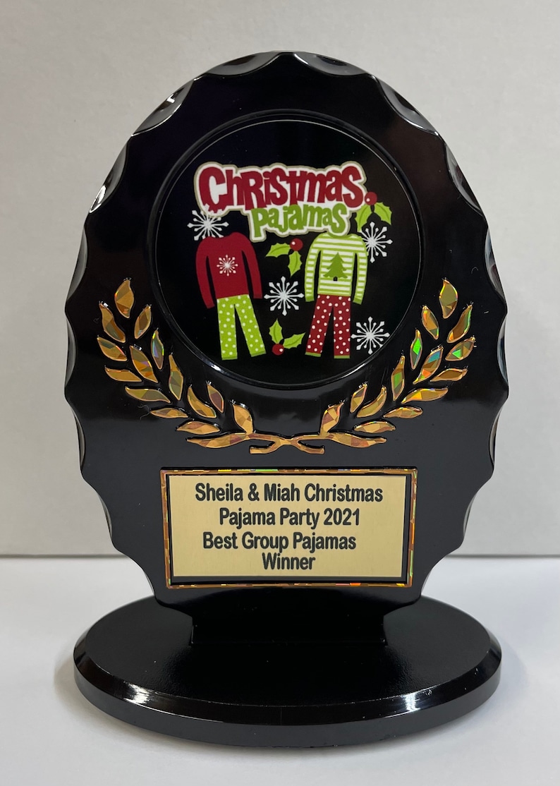 May include: Black and gold award plaque with a Christmas pajama party theme. The plaque features a circular image of two people wearing Christmas pajamas. The text on the plaque reads "Sheila & Miah Christmas Pajama Party 2021 Best Group Pajamas Winner".