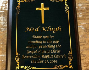 May include: A dark brown wooden plaque with a gold border and a gold cross. The plaque has a black background with gold lettering that reads "Ned Klugh Thank you for standing in the gap and for preaching the Gospel of Jesus Christ Beaverdam Baptist Church October 27, 2019".