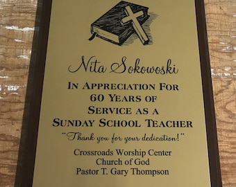 May include: A gold plaque with a brown wooden frame, honoring Nita Sokowoski for 60 years of service as a Sunday School Teacher at Crossroads Worship Center Church of God. The plaque features a black and white illustration of a closed book with a cross on top.