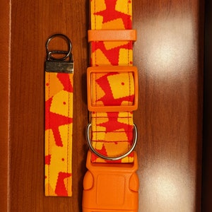 Op de afbeelding: Oranje en rode hondenhalsband en riem met een patroon van gele vierkanten. De halsband heeft een plastic gesp en een metalen D-ring.