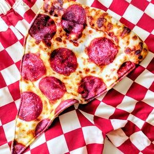 Puede incluir: Una almohada con forma de porción de pizza con pepperoni descansa sobre una tela a cuadros rojos y blancos. La pizza tiene una corteza dorada, queso blanco y rodajas redondas de pepperoni. La tela tiene una estética de restaurante clásico.