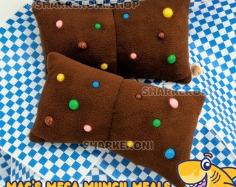 Puede incluir: Dos almohadas de felpa marrones con forma de barras de chocolate con coloridos sprinkles de caramelo. Las almohadas están sobre un fondo a cuadros azul y blanco. El texto "Mac's Mega Munch Meals" y "Only Available From Sharkeroni" está debajo de las almohadas.