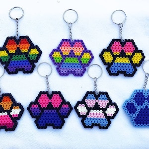 May include: Seven colorful beaded paw print keychains. The keychains are made with rainbow, purple, blue, pink, and white beads. The keychains are attached to silver key rings.