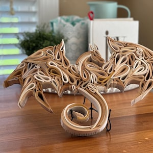 May include: A dragon sculpture made from pages of a book. The dragon is curled up with its wings spread out. The sculpture is made of paper and is painted in shades of brown and white.