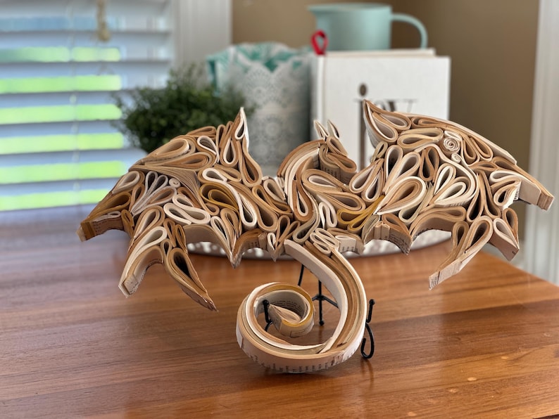 May include: A dragon sculpture made from pages of a book. The dragon is curled up with its wings spread out. The sculpture is made of paper and is painted in shades of brown and white.
