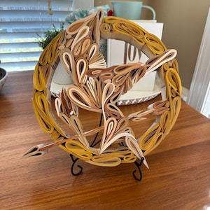 May include: A layered paper sculpture of a circular emblem with a bird and arrow design. The sculpture features shades of yellow, beige, and brown, and is mounted on a black metal stand. The artwork is displayed on a wooden table.